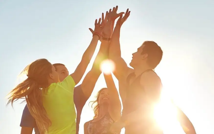 A group of athletes putting their hands together in a high-five huddle against a bright sunset background.