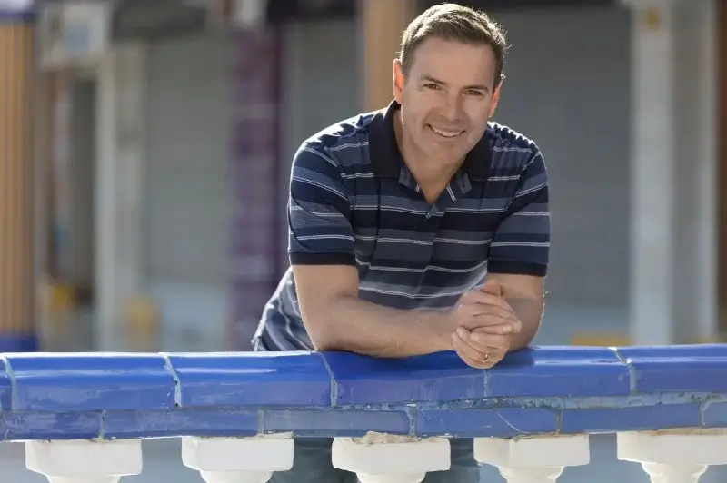 A smiling middle-aged man in a striped polo shirt leaning on a bright blue tiled railing outdoors.