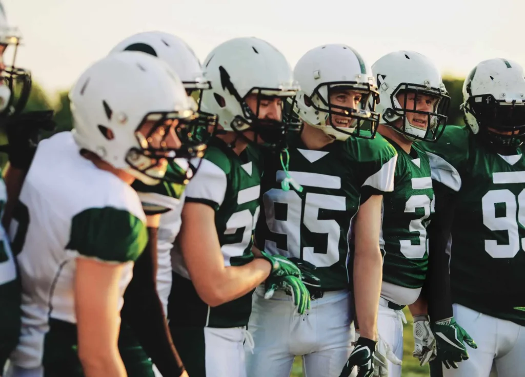 A group of American football players in green and white uniforms standing together in a team huddle on a field, wearing helmets and gloves.