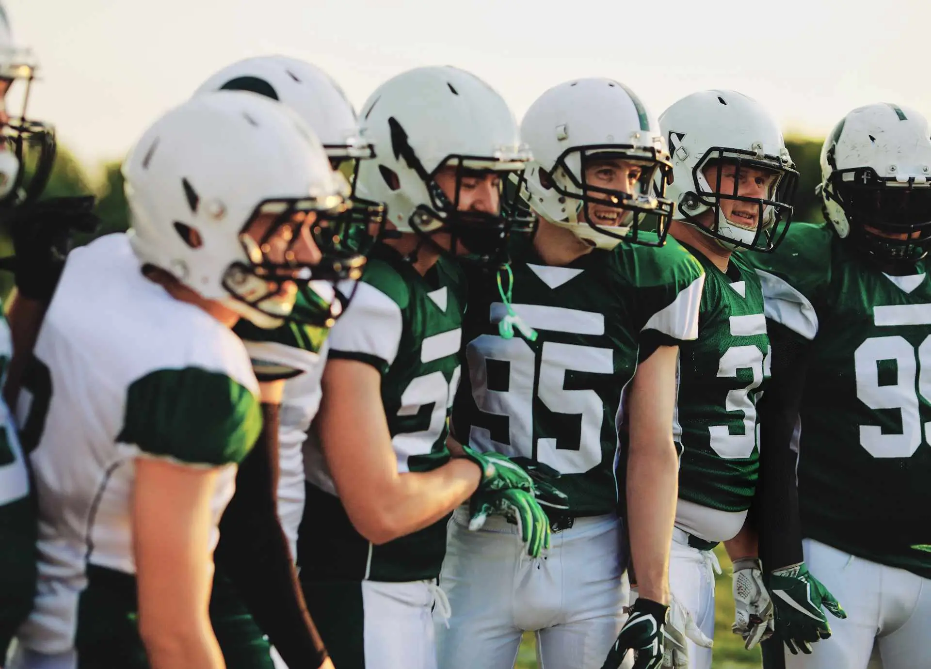 A group of American football players in green and white uniforms standing together in a team huddle on a field, wearing helmets and gloves.