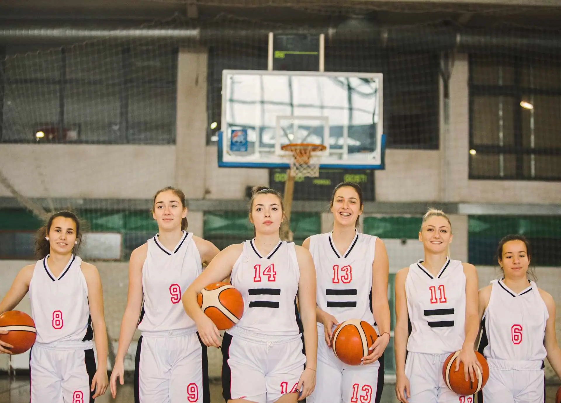 A group of six female basketball players in white uniforms standing in a line on an indoor court, holding basketballs.