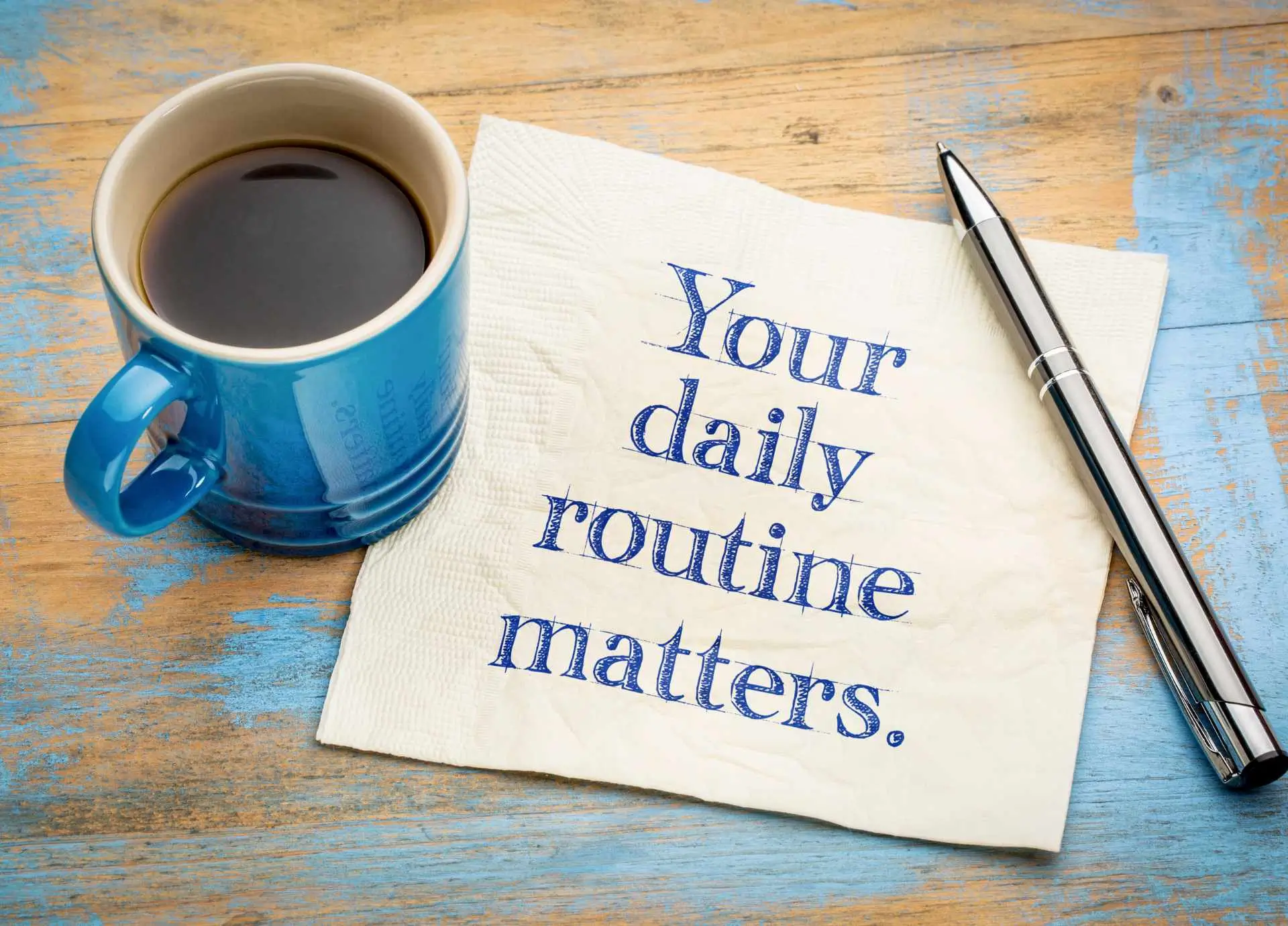 A blue coffee mug, a silver pen, and a white napkin with the text "Your daily routine matters" resting on a rustic wooden table.