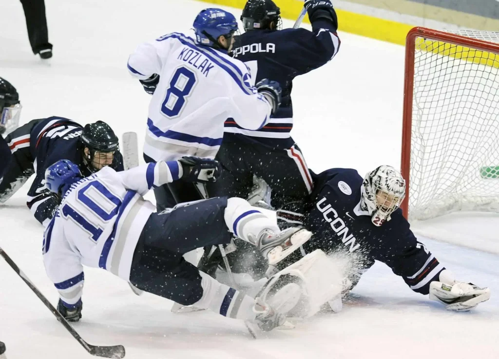 An ice hockey player in a white jersey slides on the ice near the goal while the UConn goalkeeper attempts a save.