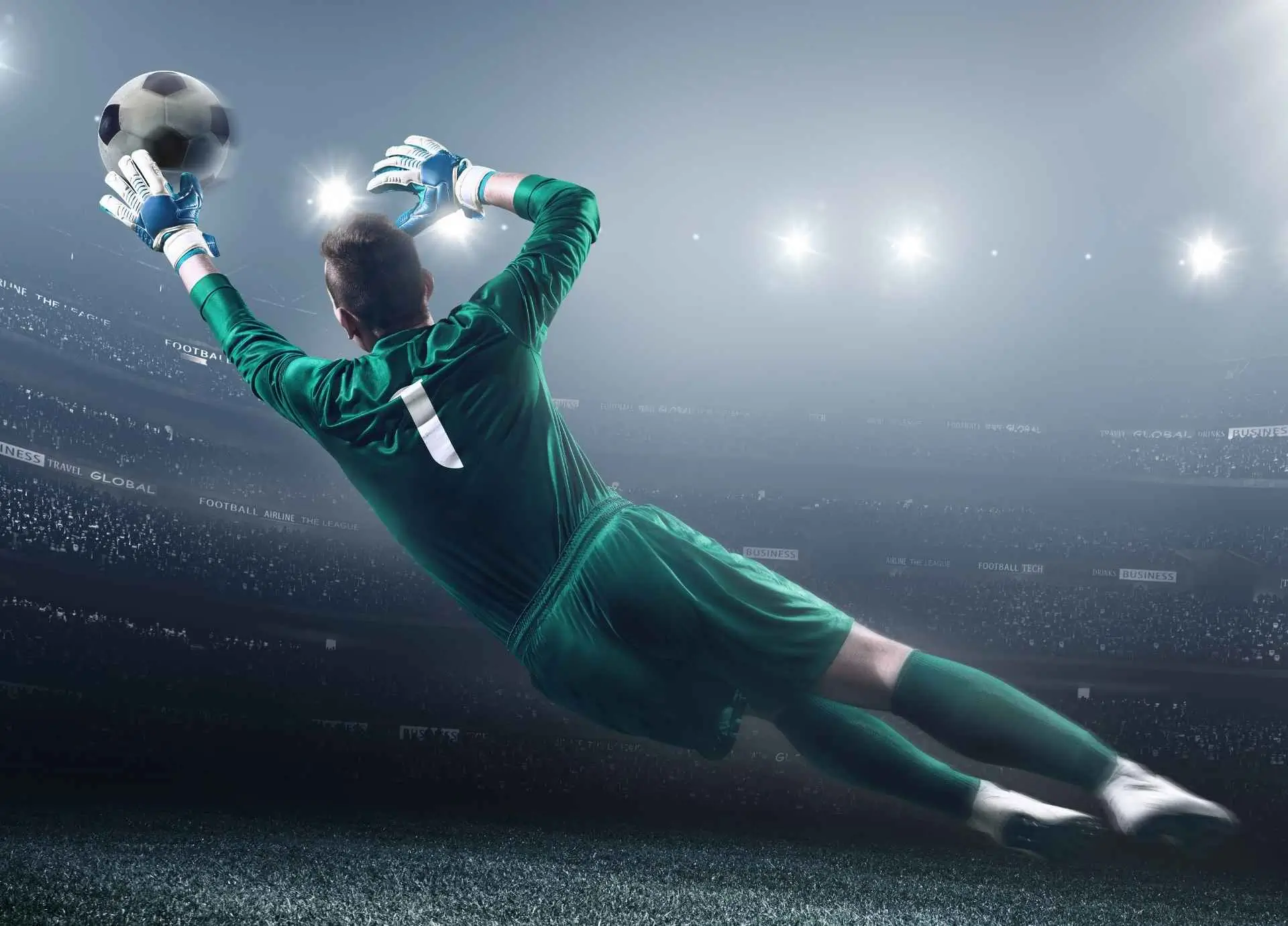 A professional soccer goalkeeper in a green jersey with the number 1 diving horizontally to catch a ball in a brightly lit stadium.