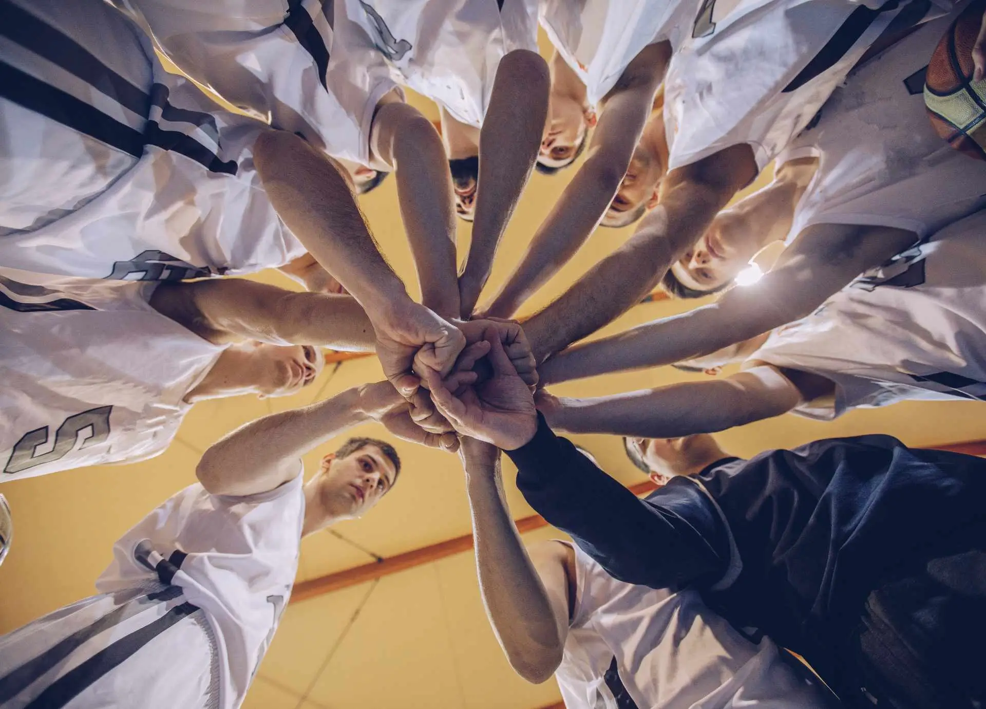 Basketball team standing in a tight circle with hands joined together in a gesture of unity and teamwork on an indoor court.