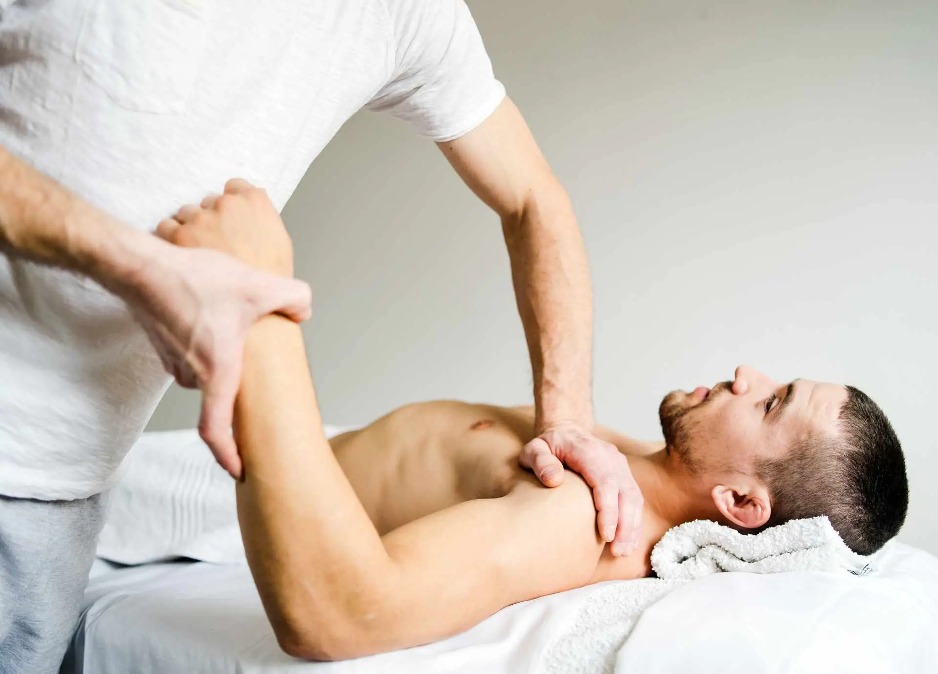 A physical therapist in a white t-shirt performs a therapeutic shoulder and arm manipulation on a male client lying on a treatment table.