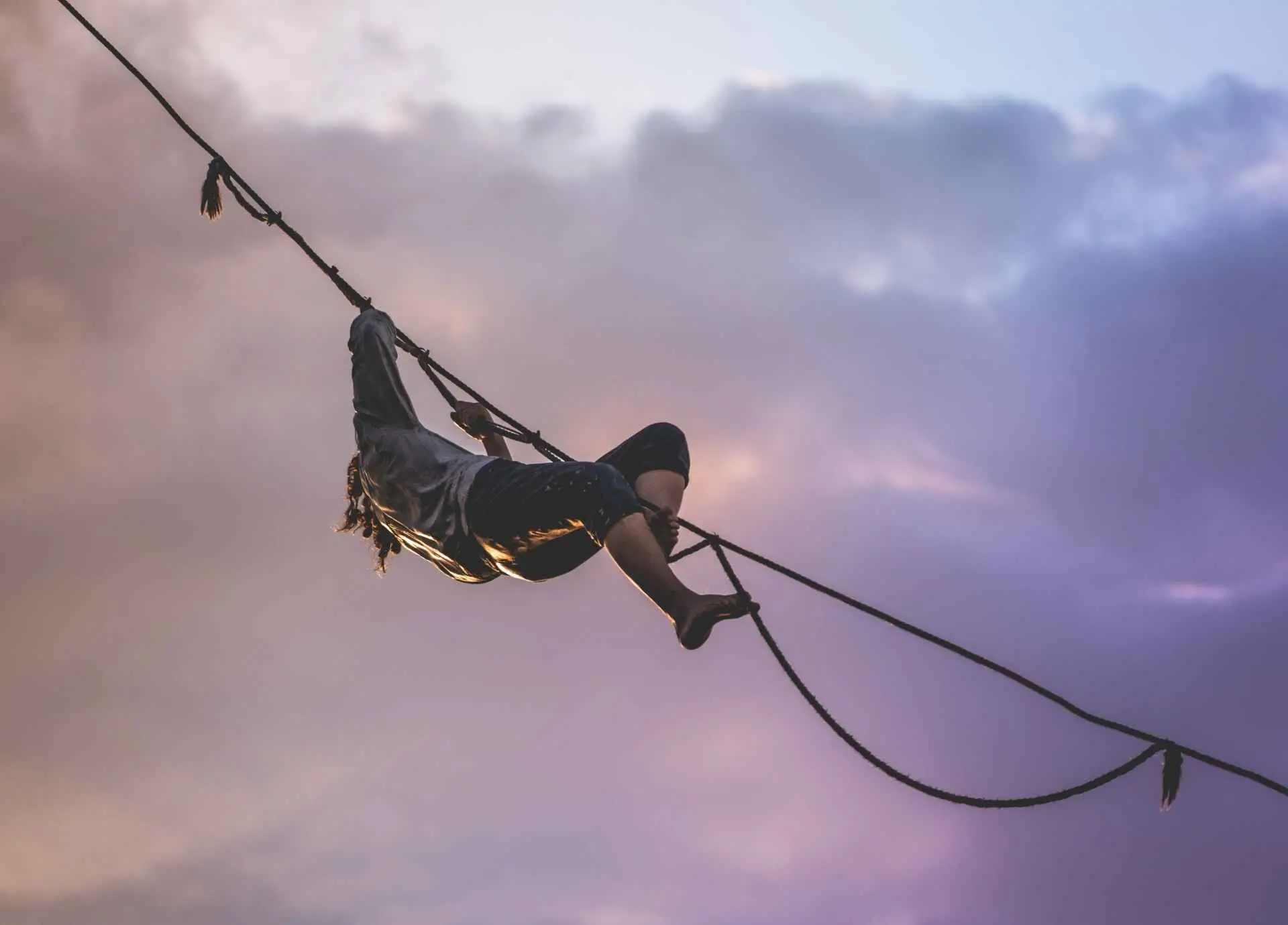 A person balancing on a highline rope against a dramatic, cloudy twilight sky.