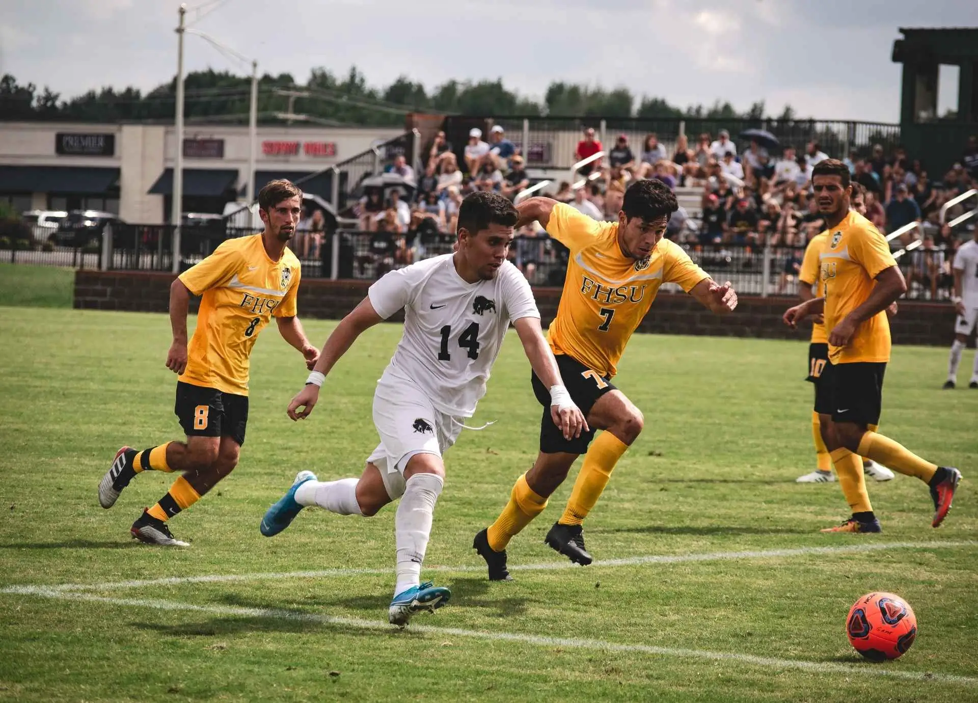 Soccer players in white and yellow jerseys competing for the ball on a sunny grass field during an intense match.