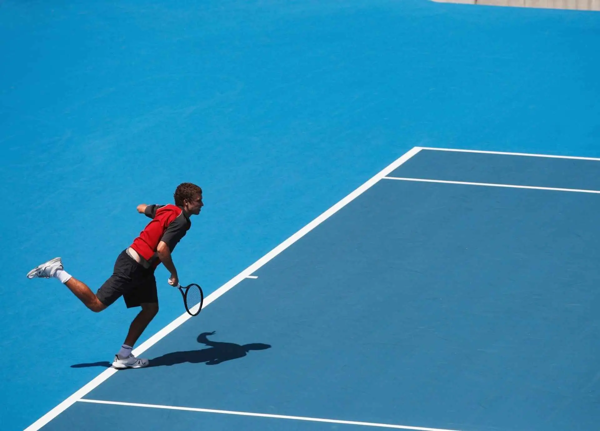 A tennis player in a red and black shirt executing a shot on a vibrant blue hard court.