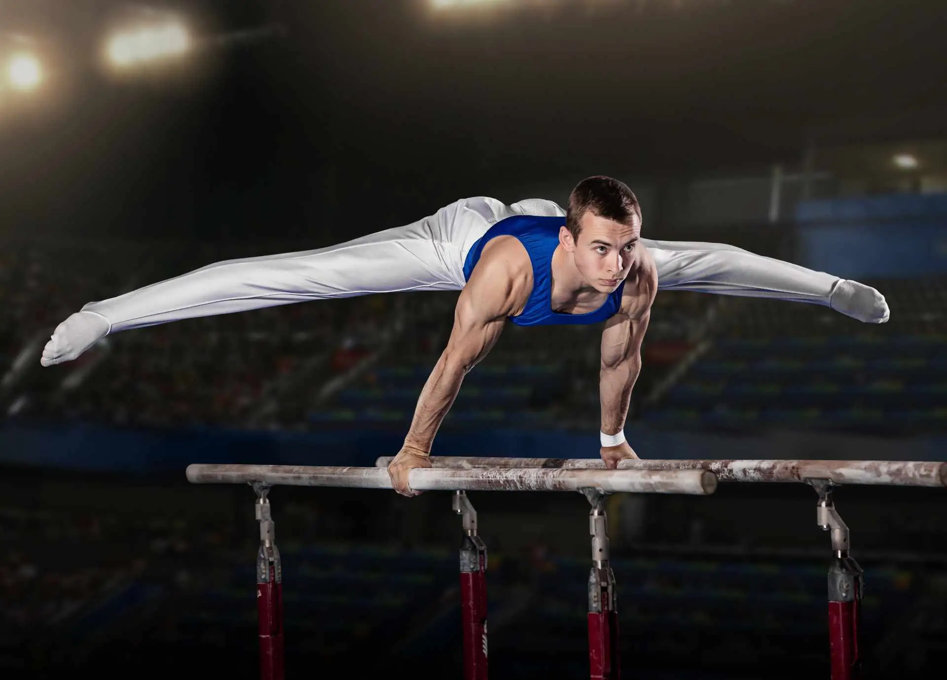 A male gymnast in a blue leotard performing a straddle position on parallel bars inside a professional arena.