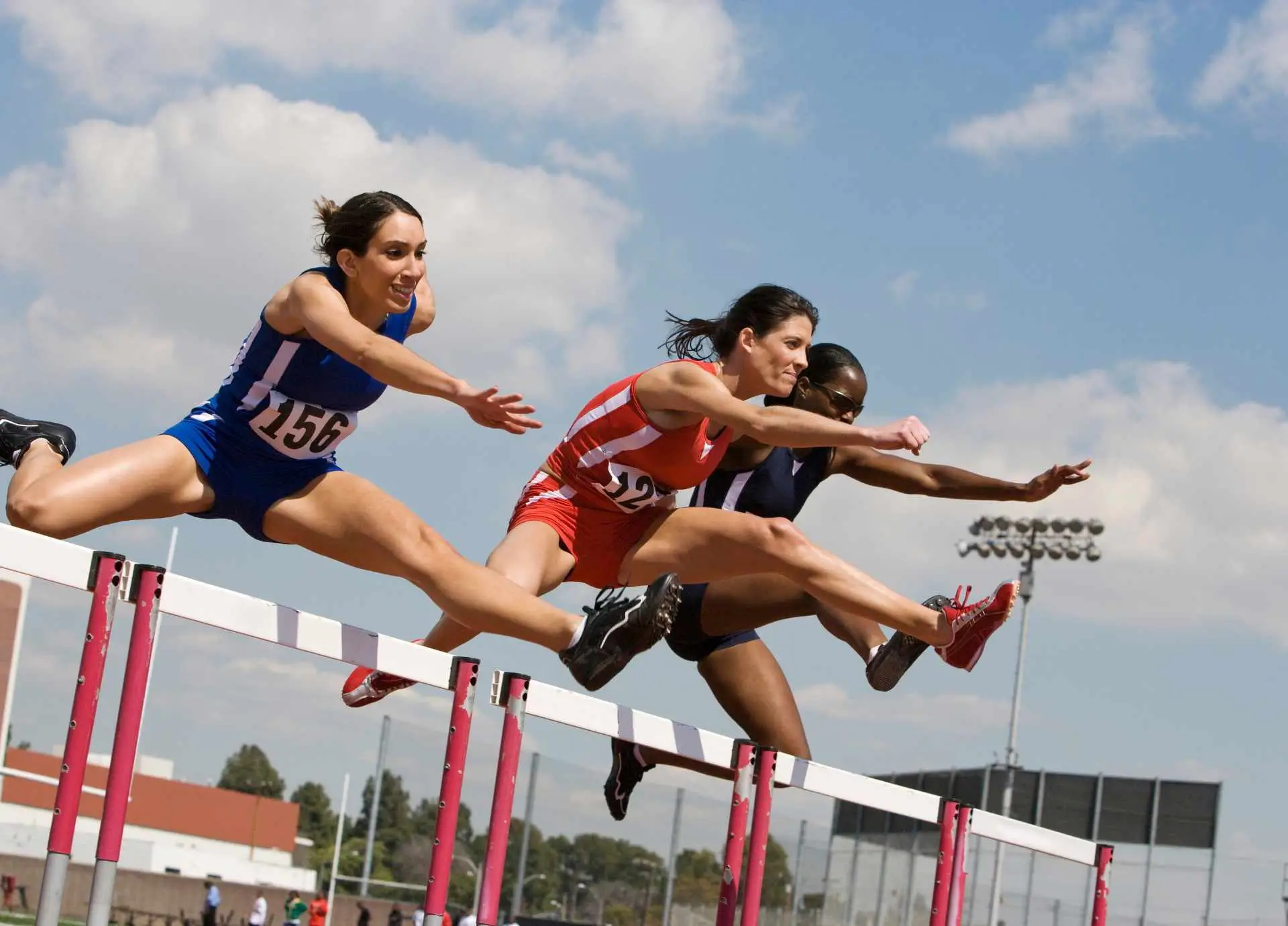Three female athletes in athletic uniforms jumping over hurdles simultaneously during a track and field competition.