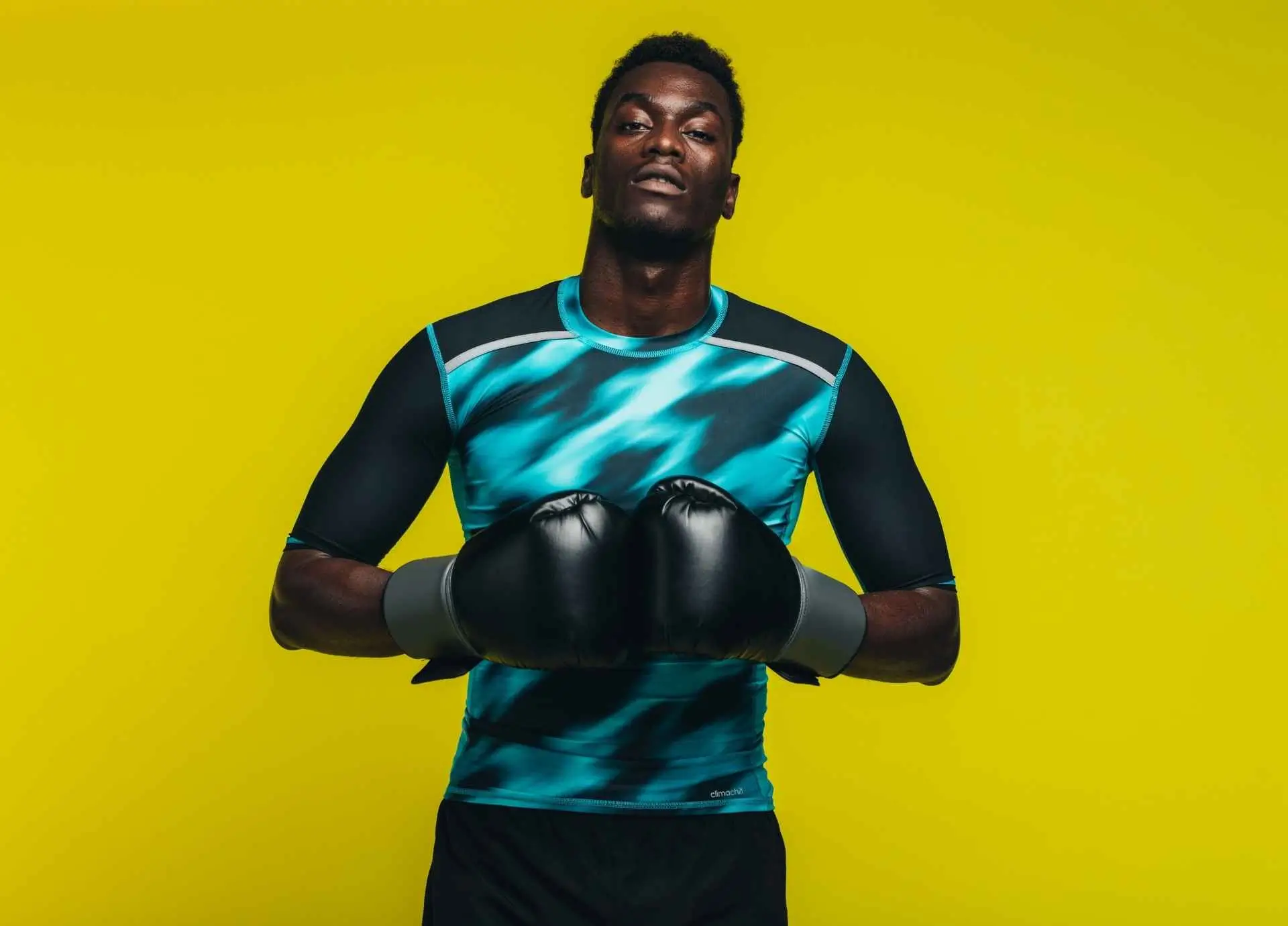A focused man wearing black boxing gloves and a patterned athletic compression shirt poses confidently against a bright yellow background.