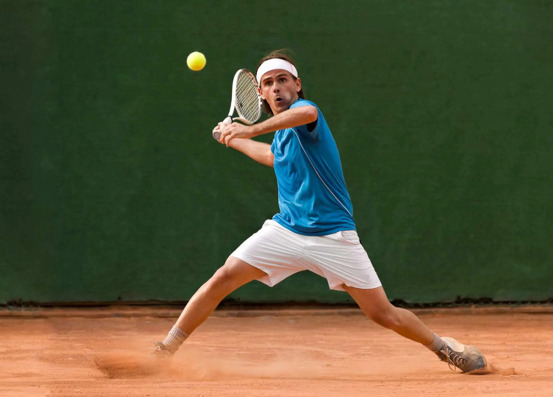 A male tennis player in a blue shirt executing a powerful forehand stroke on a clay court.