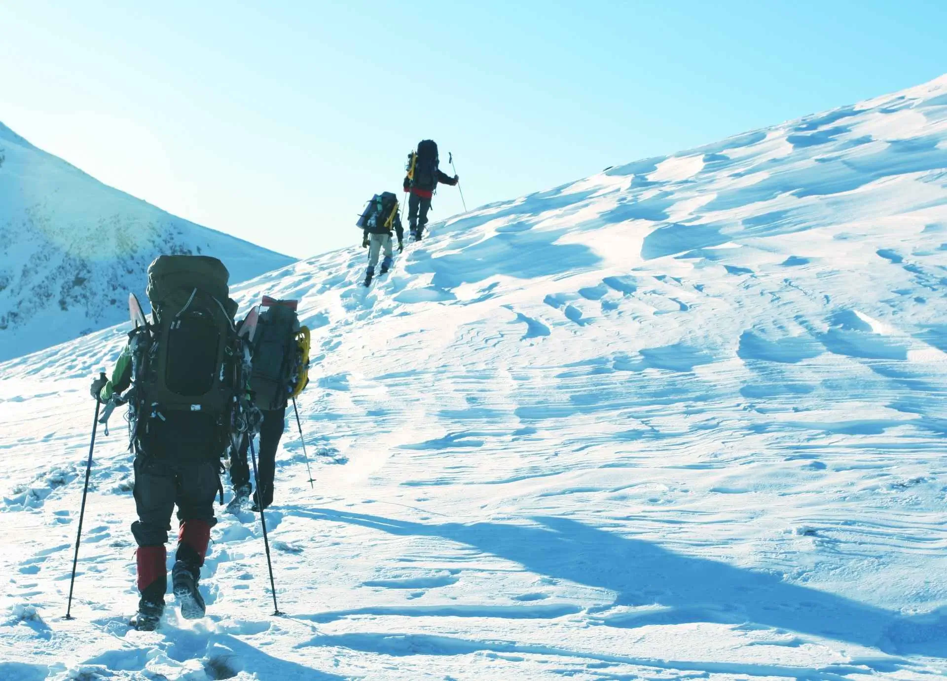 A group of three hikers with heavy backpacks and trekking poles walking up a steep, snow-covered mountain slope under a clear blue sky.