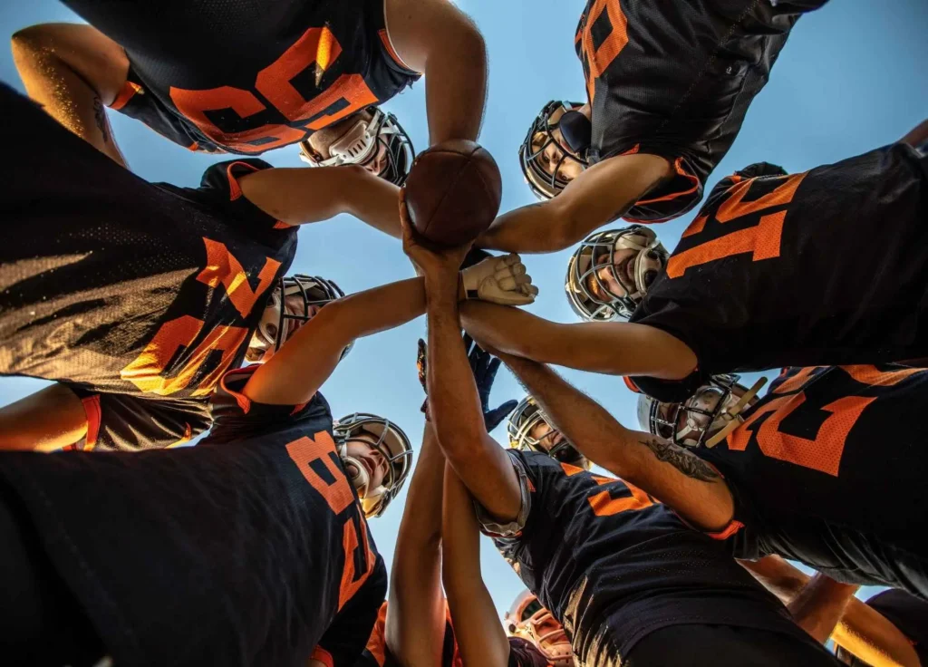 A low-angle shot of a football team in black and orange jerseys gathered in a huddle with their hands joined on a ball against a clear sky.