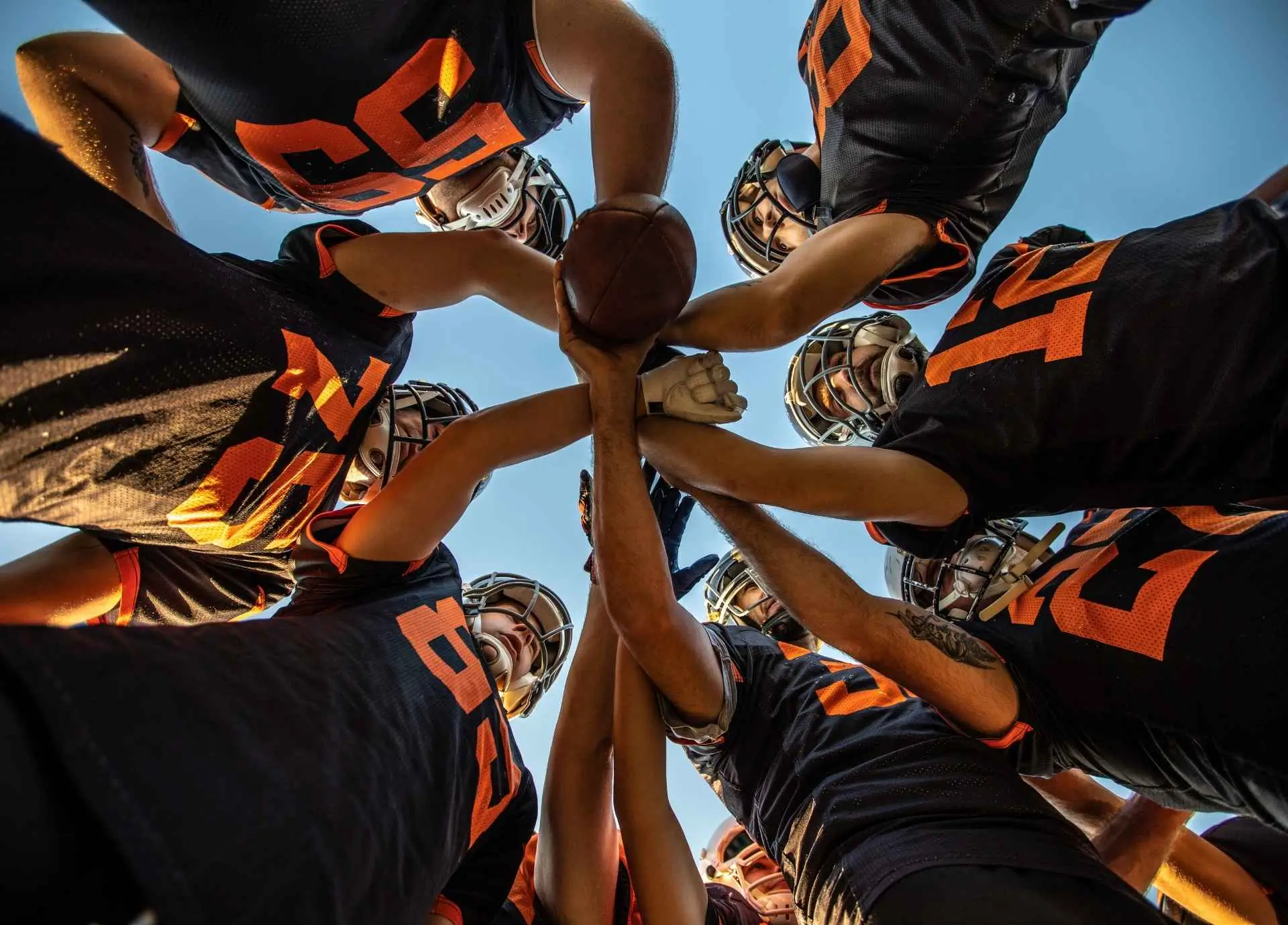 A low-angle shot of a football team in black and orange jerseys gathered in a huddle with their hands joined on a ball against a clear sky.